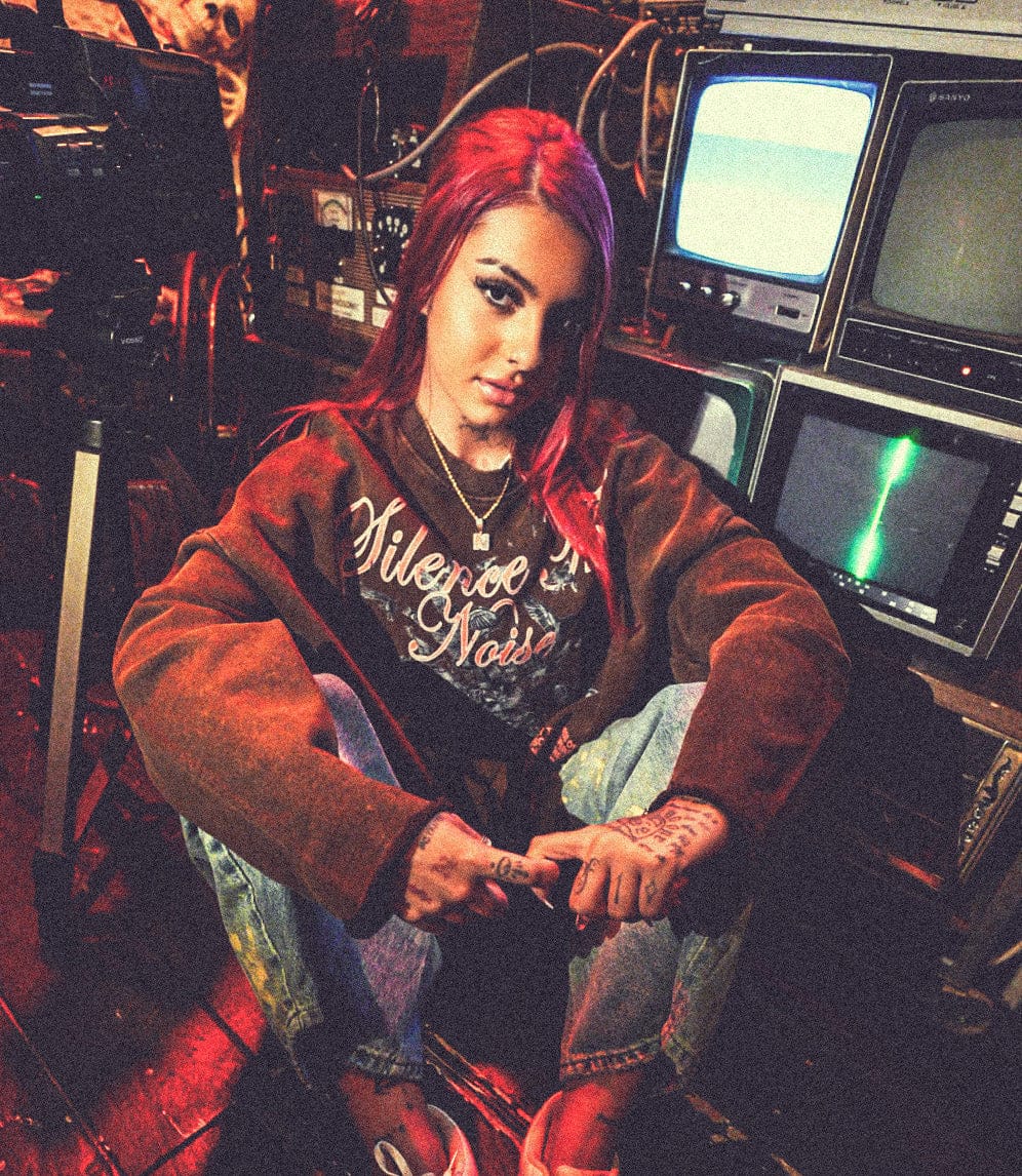 Model wearing a brown graphic hoodie and jeans, seated in an industrial room with vintage television screens